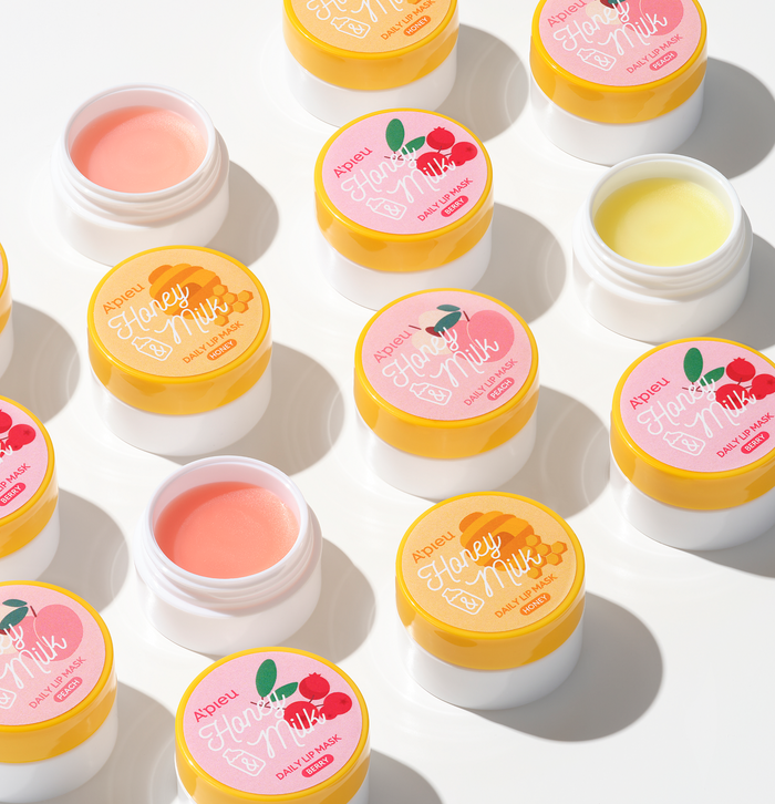 An overhead flat lay of multiple jars of A'pieu Honey Milk Daily Lip Mask on a white background, featuring three variations: Honey (yellow label with a honeycomb), Berry (pink label with raspberries), and a third variant (light pink label). Some jars are open, showing the glossy, moisturizing texture of the Korean skincare lip treatment.