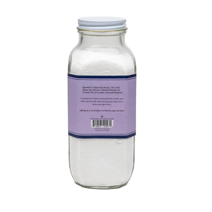 A clear glass jar with a purple label reveals "BAUDELAIRE - SLEEP BATH SELTZER." The 18 oz of white powder inside offers a handmade, therapeutic experience with its effervescent formula and natural essential oils.