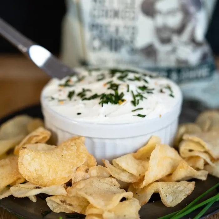 A green 5oz bag of CAROLINA KETTLE - CREAM CHEESE AND CHIVE CHIPS from 1 IN 6 SNACKS offers a gluten-free, savoury snack in every bite, with Sir Walter Raleigh holding a chip featured on the packaging.