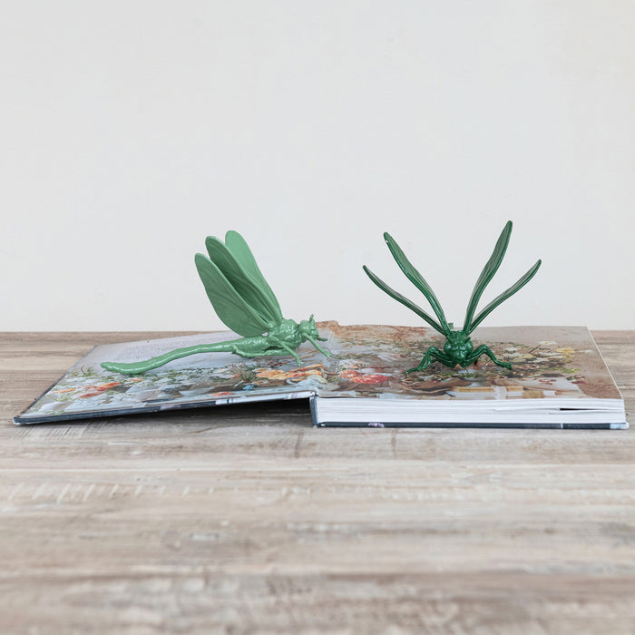 A pop-up book on a wooden surface showcases two large green CAST IRON DRAGONFLY sculptures by CREATIVE COOP, emerging from its pages against a plain light background.