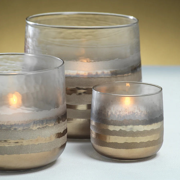 The ZODAX GOLD SMOKE VOTIVE HOLDER features a clear glass design accented with metallic gold and silver horizontal bands that fade delicately toward the top, creating a subtle metallic glow. Ideal as a smoke votive holder, it adds an ambient warmth to any space. Set against a white background.