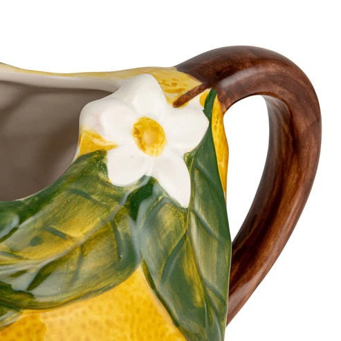 The HAND-PAINTED STONEWARE LEMON-SHAPED PITCHER by CREATIVE COOP, featuring a ceramic lemon form adorned with delicate floral and leafy details near the handle, is displayed beside a folded cloth, a halved grapefruit, and a yellow dish on a light background.
