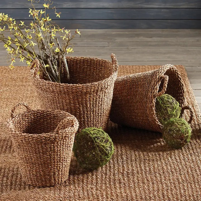 The NAPA HOME AND GARDEN Seagrass Tapered Basket with Handles and Cuff, Small, sits on a textured rug before a white paneled wall, alongside two other baskets, with part of a chair and a colorful painting in the background.