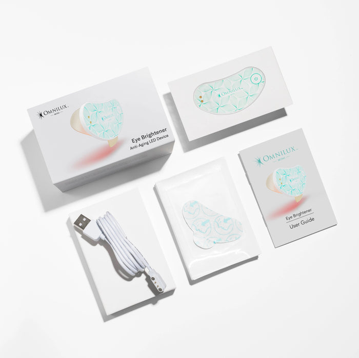 White wearable eye mask with a green leaf pattern, labeled “OMNILUX – EYE BRIGHTENER KIT,” featuring a power button and two small metal contacts, softly glowing with a red light underneath to deliver targeted red light therapy for brightening the eye area against a plain gray background.