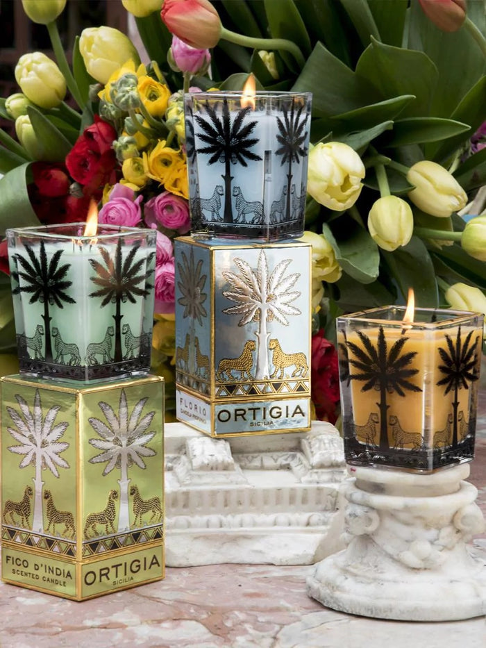The ORTIGIA SICILIA - FLORIO LARGE SQUARE CANDLE features pale blue wax in a decorative gattopardo-printed jar, with both the candle and its box adorned with gold and black palm trees and leopards against a pale blue background—an elegant tribute to Sicilian spring flowers.