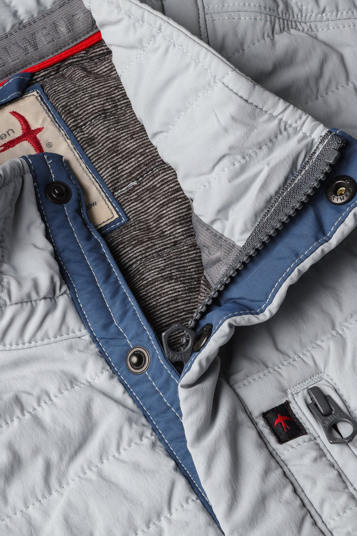 A person models the RELWEN - WINDZIP VEST IN GHOST GRAY over a gray sweater, plaid shirt, and dark blue jeans, standing with one hand in their pocket against a white background for extra layered warmth.