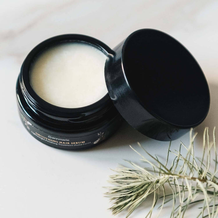 Black hair serum jar with open lid on a white surface with green leaves.