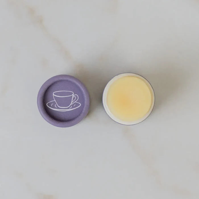 Round container with purple lid featuring a tea cup design, filled with a yellowish substance on a light marble background.
