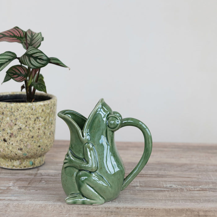 The CREATIVE COOP STONEWARE FROG PITCHER with a green reactive glaze sits on a wooden surface beside a potted variegated plant, against a plain light background.