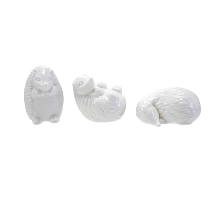 Six CREATIVE COOP WHITE STONEWARE HEDGEHOG figurines in different poses are arranged on and around three whitewashed cylindrical wooden pedestals, perfect for charming farmhouse decor. The background is plain white.