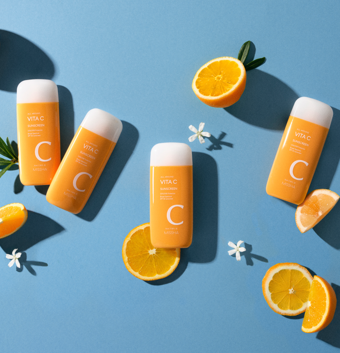 Four orange sunscreen bottles are arranged on a blue background, surrounded by fresh orange slices and white flowers.