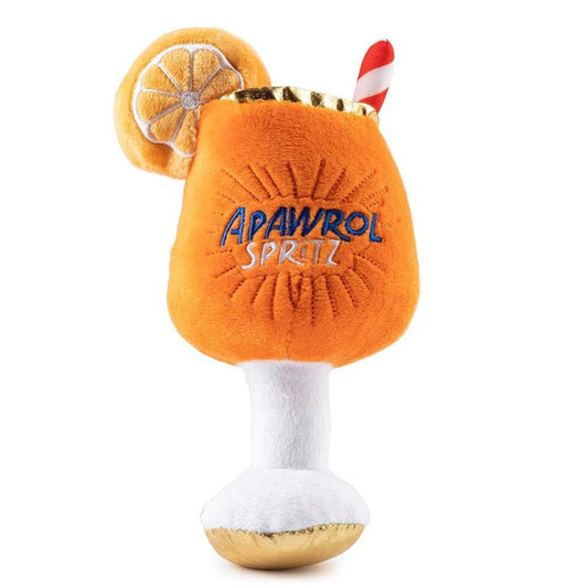 A fluffy Pomeranian dog sits on a rug outdoors next to the HAUTE DIGGITY DOG APAWROL SPRITZ DOG TOY, a squeaky toy shaped like an orange cocktail with a straw and lemon slice.
