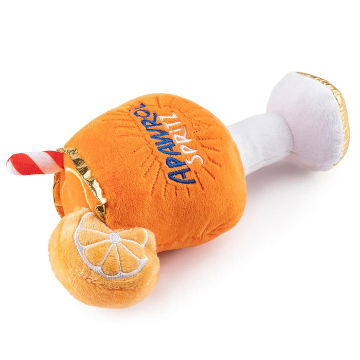 A fluffy Pomeranian dog sits on a rug outdoors next to the HAUTE DIGGITY DOG APAWROL SPRITZ DOG TOY, a squeaky toy shaped like an orange cocktail with a straw and lemon slice.