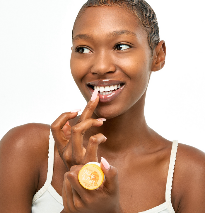smiling model with short, natural hair applies the A'pieu Honey Milk Daily Lip Mask in the Honey flavor to her lips with her finger. The gold-capped jar is visible in her hand. Her lips have a glossy, hydrated look, emphasizing the product's moisturizing and softening benefits.