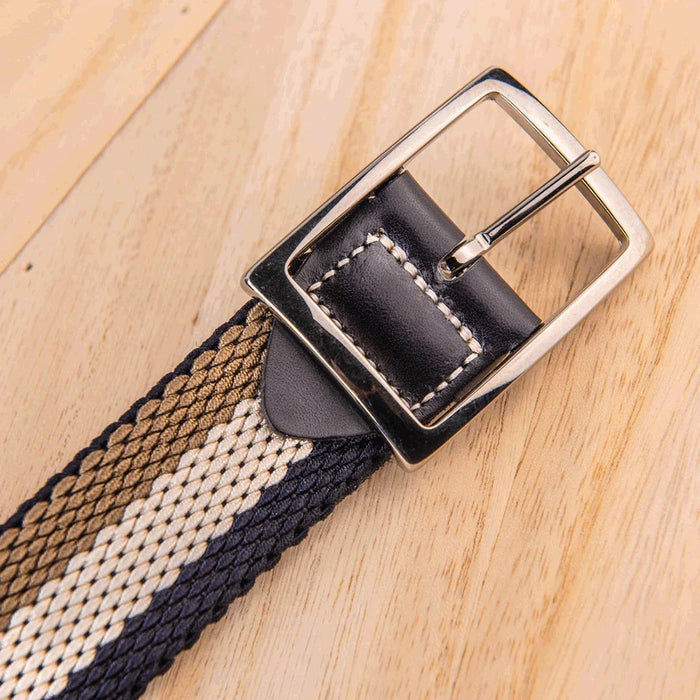 A close-up of the ATHISON Reversible Stretch Belt in navy, khaki, and white stripes with a silver buckle and navy leather details, shown on a light wooden surface; reverses to solid navy.