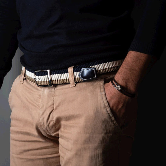 A close-up of the ATHISON Reversible Stretch Belt in navy, khaki, and white stripes with a silver buckle and navy leather details, shown on a light wooden surface; reverses to solid navy.