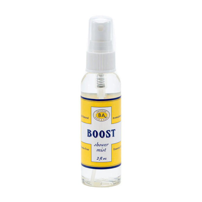 A collection of BAUDELAIRE's "Boost" skincare products, including bath salts, cleansing oil, and a peppermint-infused BAUDELAIRE - BOOST SHOWER MIST in a spray bottle, is displayed on a shelf alongside a tall yellow vase with green and brown stems.