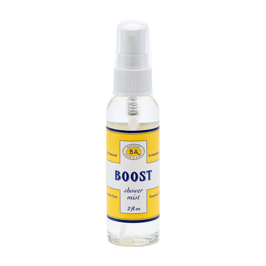 A collection of BAUDELAIRE's "Boost" skincare products, including bath salts, cleansing oil, and a peppermint-infused BAUDELAIRE - BOOST SHOWER MIST in a spray bottle, is displayed on a shelf alongside a tall yellow vase with green and brown stems.