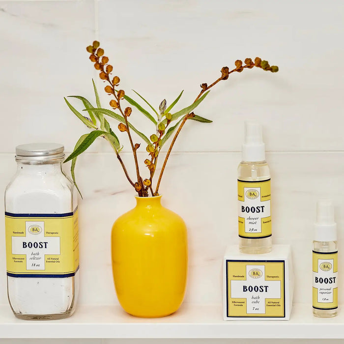 A collection of BAUDELAIRE's "Boost" skincare products, including bath salts, cleansing oil, and a peppermint-infused BAUDELAIRE - BOOST SHOWER MIST in a spray bottle, is displayed on a shelf alongside a tall yellow vase with green and brown stems.