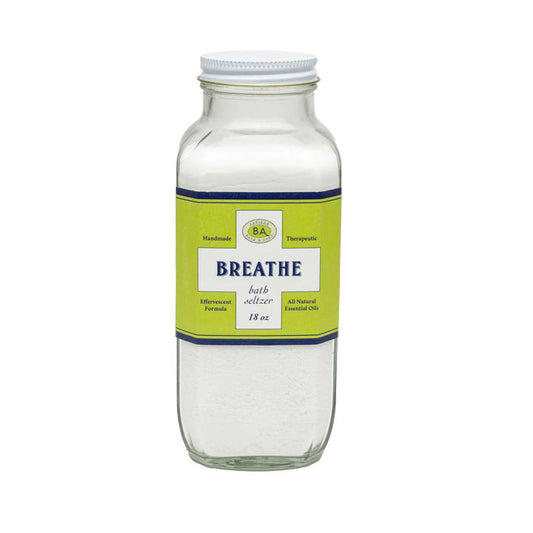 A shelf displays BAUDELAIRE Breathe Bath Seltzer alongside essential oils, a quirky face-shaped pot with a plant, and a wooden brush. The background showcases a geometric pattern.