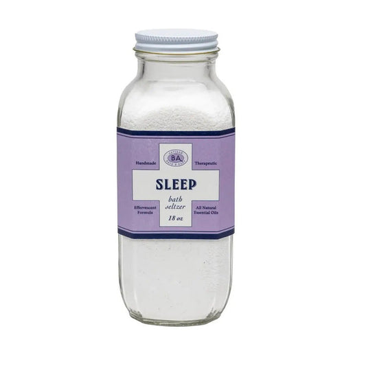 A clear glass jar labeled "BAUDELAIRE - SLEEP BATH SELTZER" features a purple label with ingredients, a barcode, and essential oils. Filled with granular white bath salts, its decorative border suggests instructions for a calming soak.
