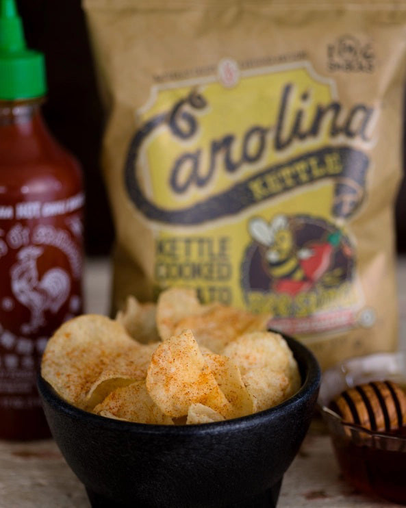 A 5oz bag of CAROLINA KETTLE - HONEY SRIRACHA CHIPS from 1 IN 6 SNACKS comes in tan packaging with a cartoon bee holding a honey dipper and bold yellow-black text, representing the sweet and spicy chips inside.