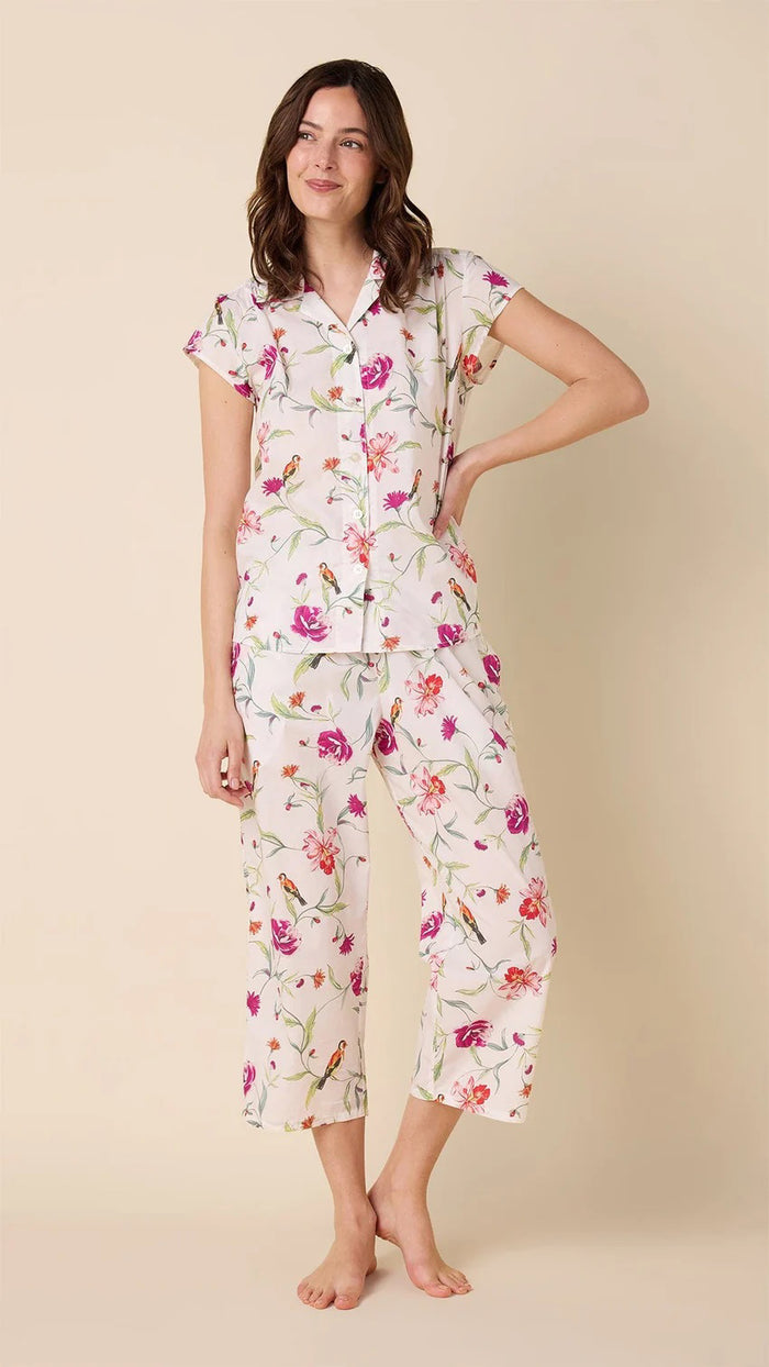 A person stands barefoot, wearing the CAT'S PAJAMAS - ROSA AND BIRD LUXE PIMA CAPRI PAJAMA SET from CAT'S PAJAMAS, featuring pink, purple, and green florals on a cream 100% cotton base with short sleeves and cropped pants. The backdrop is light beige.