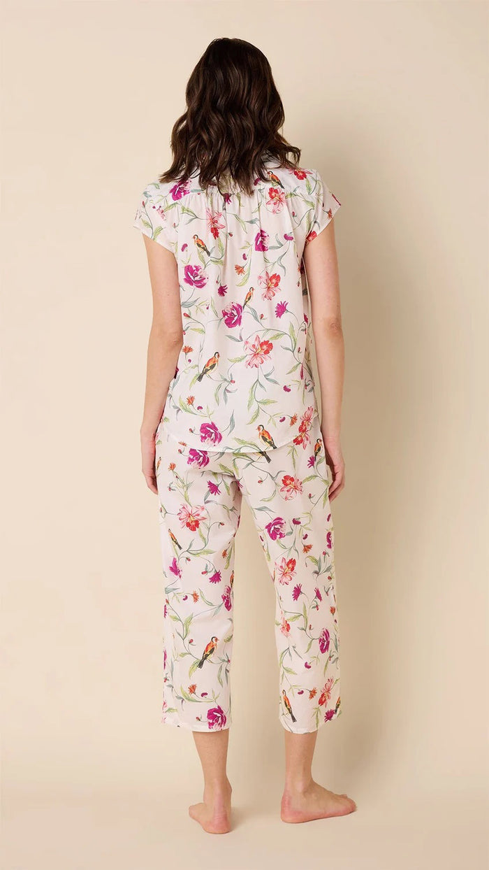 A person smiles while wearing the CAT'S PAJAMAS - ROSA AND BIRD LUXE PIMA CAPRI PAJAMA SET, featuring a floral design on 100% cotton with short sleeves and capri pants. The fabric is light in color with colorful flowers, balancing comfort and style against a plain, light background.