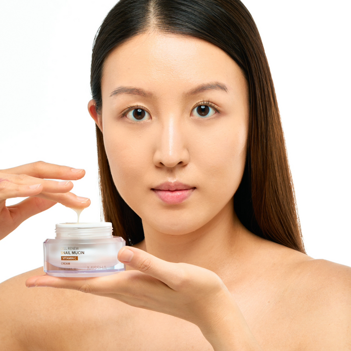 Portrait of an Asian woman holding the jar of Missha Cell Renew Snail Mucin Vitamin C Cream and scooping out product, emphasizing its role in a brightening and firming skincare routine.
