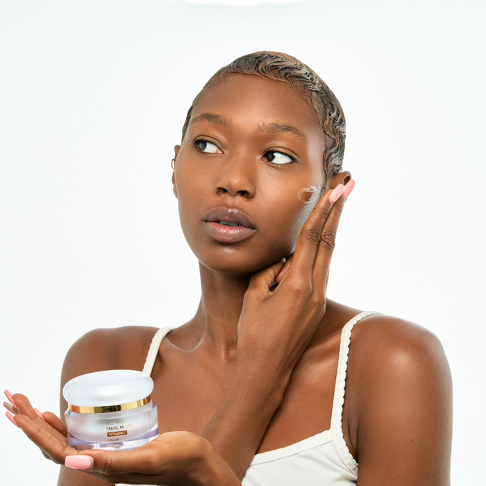 A Black model holding the jar of Missha Cell Renew Snail Mucin Vitamin C Cream while applying a small amount to her temple, highlighting the cream's benefits for hydration and uneven skin tone for melanated skin.