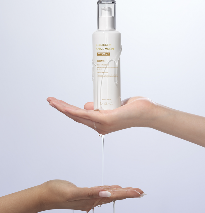A hand holding the Missha Cell Renew Snail Mucin Vitamin C Essence pump bottle over a second hand, with the serum dripping down. This shows the hydrating, lightweight formula being dispensed for daily K-Beauty routine use.