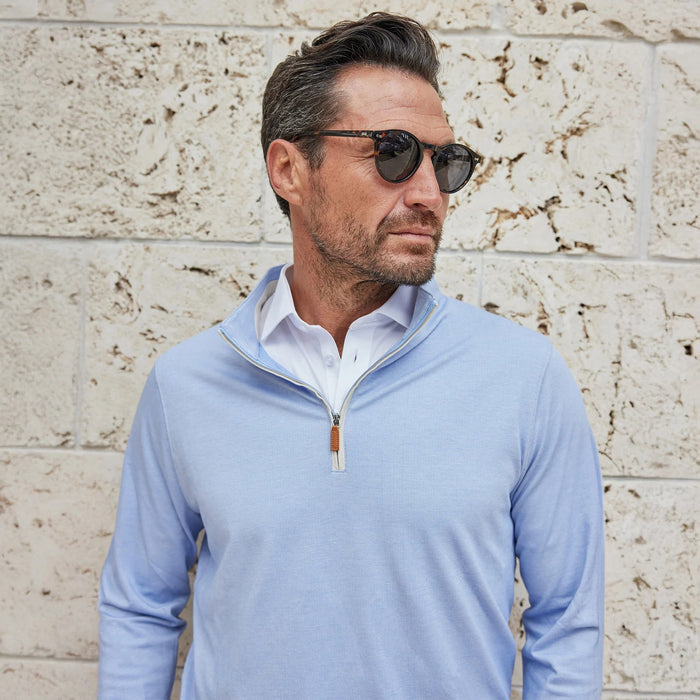 A smiling man in white pants and the COLLARS AND CO - PALM PIMA BLEND QUARTER ZIP IN BLUE holds sunglasses while walking on a sunny, palm-lined city street with shops.