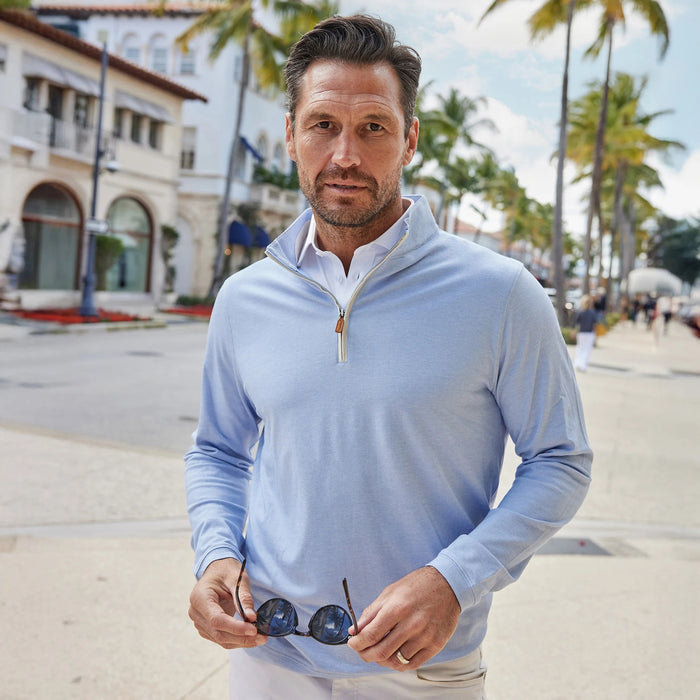 A smiling man in white pants and the COLLARS AND CO - PALM PIMA BLEND QUARTER ZIP IN BLUE holds sunglasses while walking on a sunny, palm-lined city street with shops.