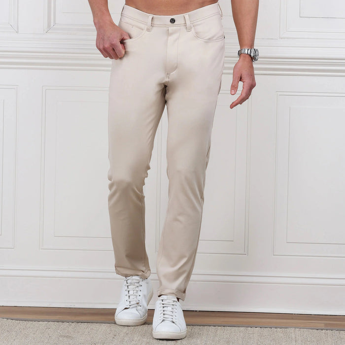 A man stands against a white paneled wall, hands in pockets, wearing COLLARS AND CO Velocity Pull-On Performance Pants in Stone with white sneakers. These wrinkle-resistant pants from COLLARS AND CO deliver effortless style; his upper body is not shown.