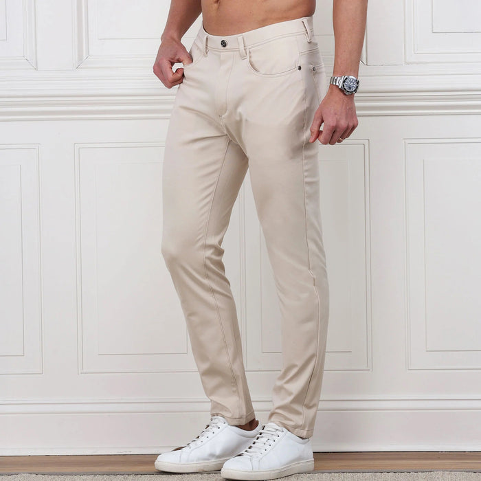 A man stands against a white paneled wall, hands in pockets, wearing COLLARS AND CO Velocity Pull-On Performance Pants in Stone with white sneakers. These wrinkle-resistant pants from COLLARS AND CO deliver effortless style; his upper body is not shown.