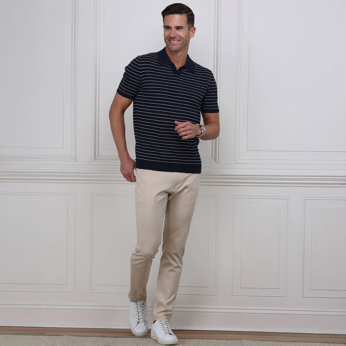 A man stands against a white paneled wall, hands in pockets, wearing COLLARS AND CO Velocity Pull-On Performance Pants in Stone with white sneakers. These wrinkle-resistant pants from COLLARS AND CO deliver effortless style; his upper body is not shown.