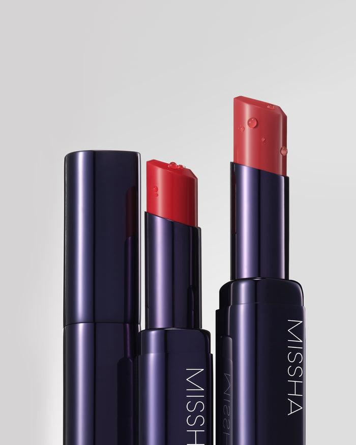 Close-up of sleek MISSHA lipsticks in deep navy tubes, showing two shades of rich coral-red lipstick with a smooth, glossy finish and visible moisture droplets on the surface.