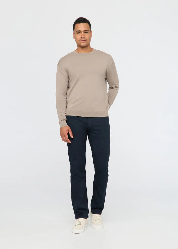 A man faces forward in a beige long-sleeve sweater, white sneakers, and DUER NO SWEAT STRAIGHT PANTS IN NAVY, set against a plain light backdrop.