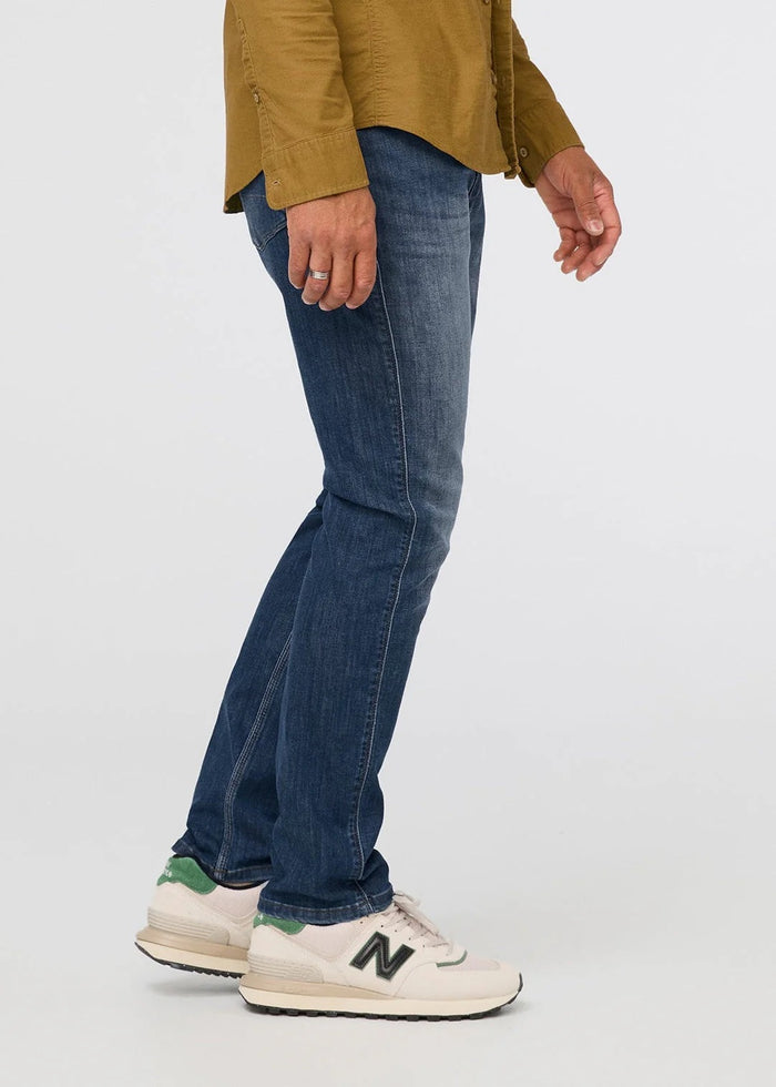 Wearing DUER’s Performance Denim Athletic Taper Pants in Galactic, paired with a mustard jacket and white shirt, a person stands against a plain light background with hands relaxed; the cropped image shows only from shoulders down.