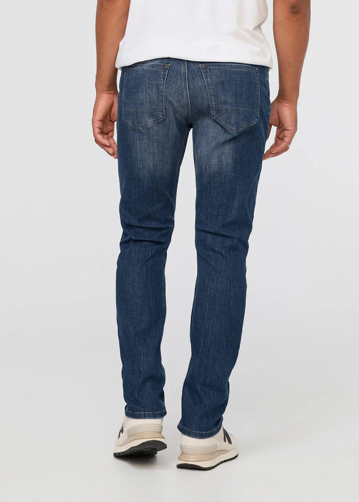 Wearing DUER’s Performance Denim Athletic Taper Pants in Galactic, paired with a mustard jacket and white shirt, a person stands against a plain light background with hands relaxed; the cropped image shows only from shoulders down.