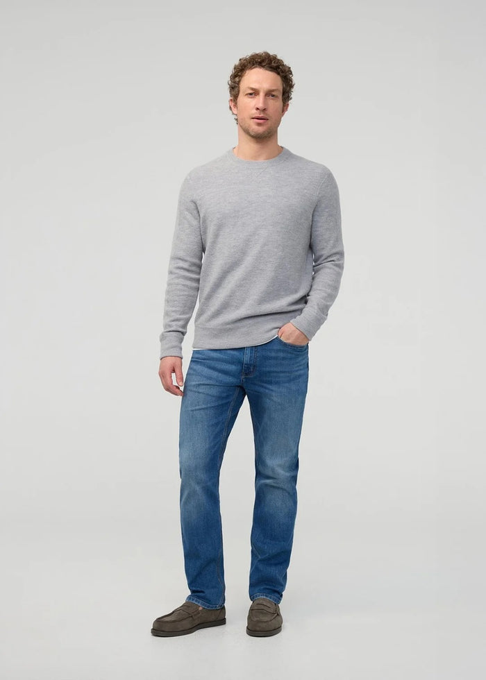 Close-up of a person wearing DUER’s PERFORMANCE DENIM+ Straight Pants in Alpine Indigo, showing relaxed hands and highlighting the texture, stitching, and stretch comfort of these blue jeans against a plain light background.
