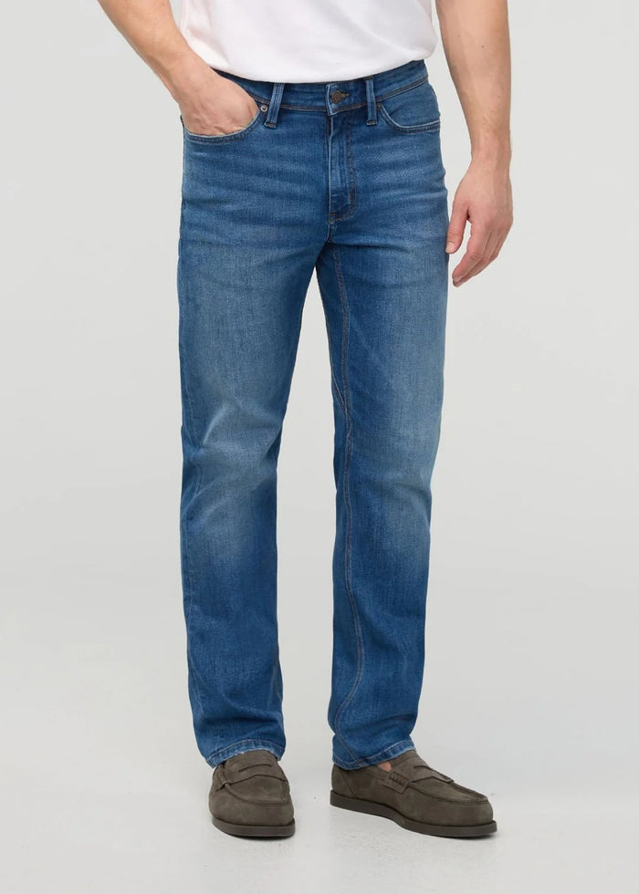 Close-up of a person wearing DUER’s PERFORMANCE DENIM+ Straight Pants in Alpine Indigo, showing relaxed hands and highlighting the texture, stitching, and stretch comfort of these blue jeans against a plain light background.