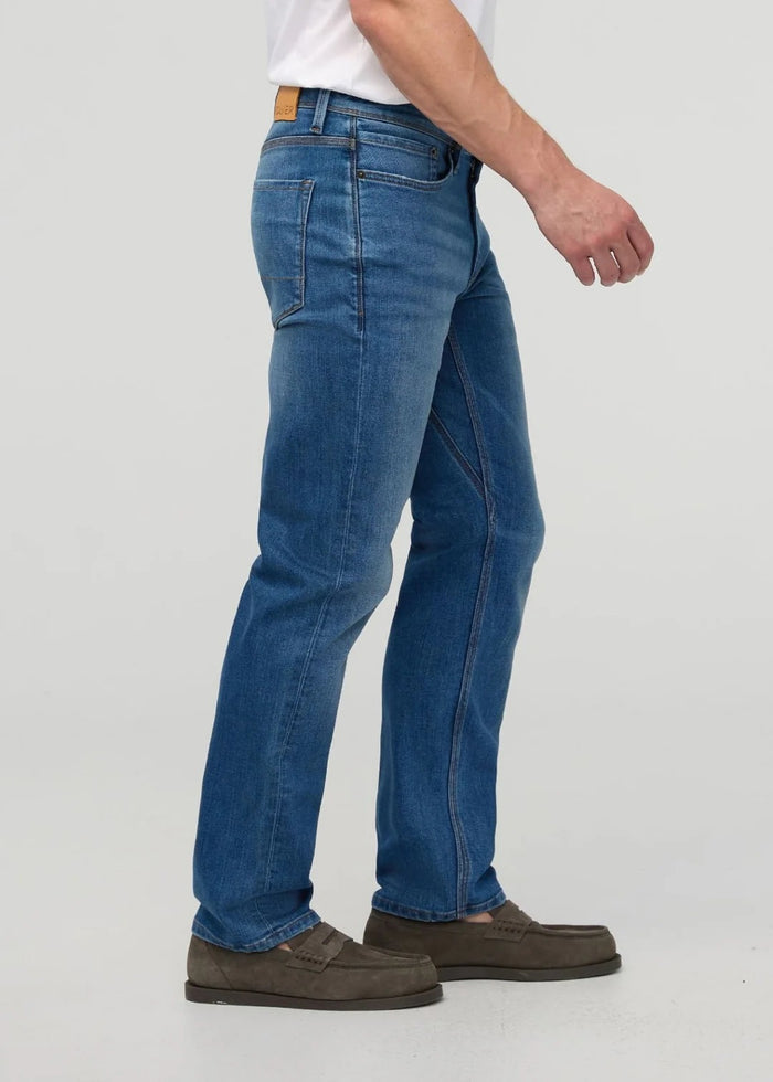 Close-up of a person wearing DUER’s PERFORMANCE DENIM+ Straight Pants in Alpine Indigo, showing relaxed hands and highlighting the texture, stitching, and stretch comfort of these blue jeans against a plain light background.