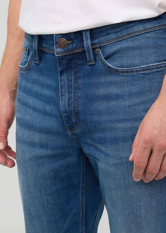 Close-up of a person wearing DUER’s PERFORMANCE DENIM+ Straight Pants in Alpine Indigo, showing relaxed hands and highlighting the texture, stitching, and stretch comfort of these blue jeans against a plain light background.