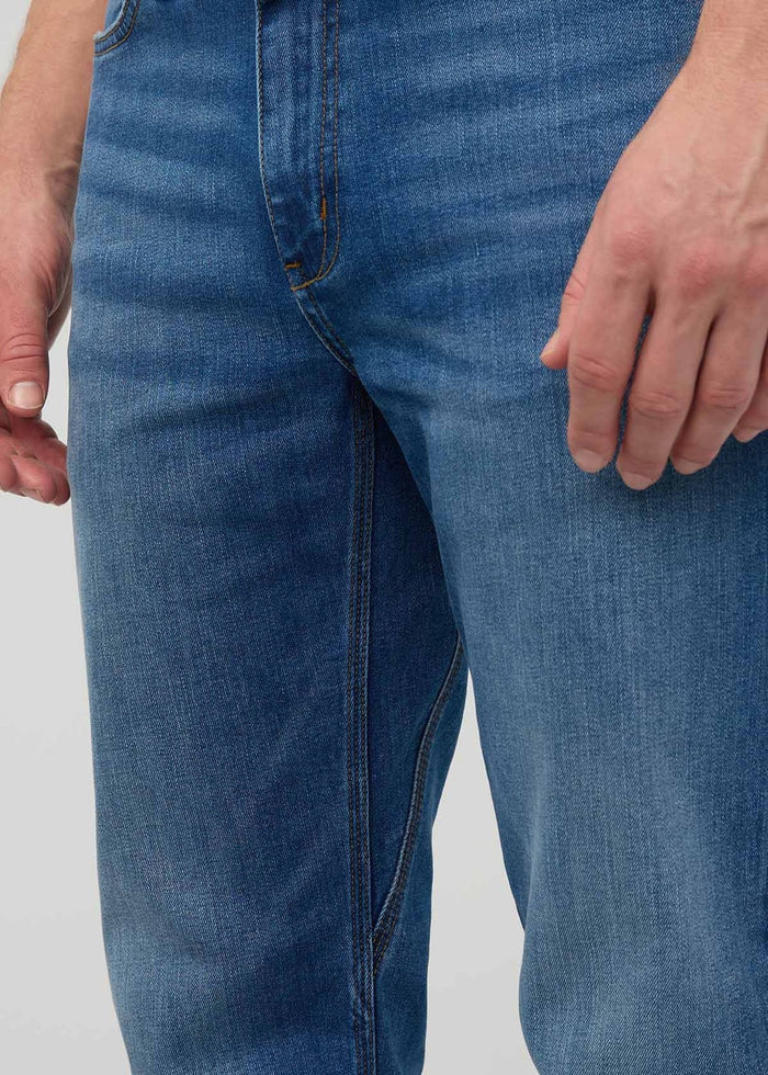 Close-up of a person wearing DUER’s PERFORMANCE DENIM+ Straight Pants in Alpine Indigo, showing relaxed hands and highlighting the texture, stitching, and stretch comfort of these blue jeans against a plain light background.