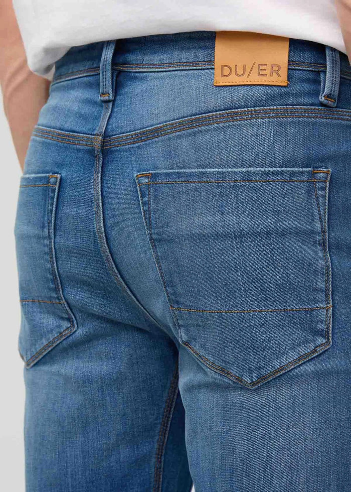 Close-up of a person wearing DUER’s PERFORMANCE DENIM+ Straight Pants in Alpine Indigo, showing relaxed hands and highlighting the texture, stitching, and stretch comfort of these blue jeans against a plain light background.