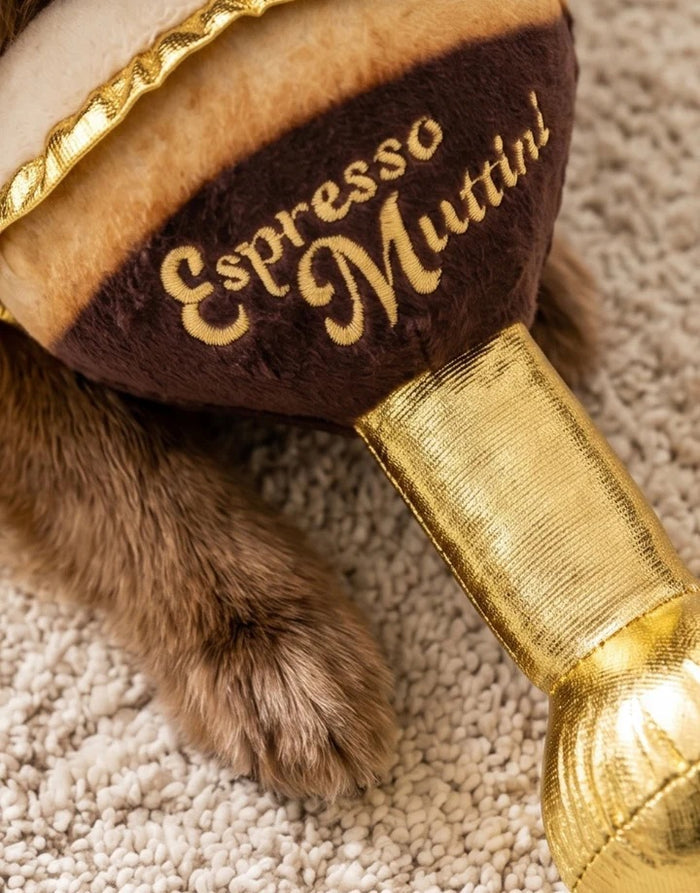 An espresso martini-shaped plush from HAUTE DIGGITY DOG, the ESPRESSO MUTTINI DOG TOY, rests on a marble table beside a coffee cup. This durable dog toy features gold, brown, and cream colors and displays “Espresso Mutini” on the side.