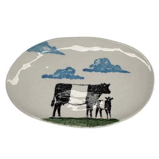 The FEARRINGTON SIGNATURE COLLECTION Beltie Oval Platter by VIETRI features black cow and calf silhouettes on green grass with blue clouds on a cream base, making it a charming gift inspired by Fearrington Village.