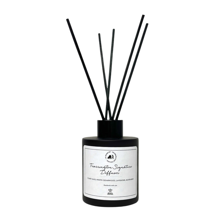 Housed in a sleek black bottle with five slender black reeds, the FEARRINGTON SIGNATURE DIFFUSER from the FEARRINGTON SIGNATURE COLLECTION by FEARRINGTON VILLAGE - HOME DECOR features a white label displaying "Fragrance Signature Diffuser" and scent notes of cedarwood, lavender, and amber. Inspired by the tranquil allure of Fearrington Village, this sophisticated reed diffuser is ideal for anyone who appreciates the calming atmosphere created by a lavender candle.