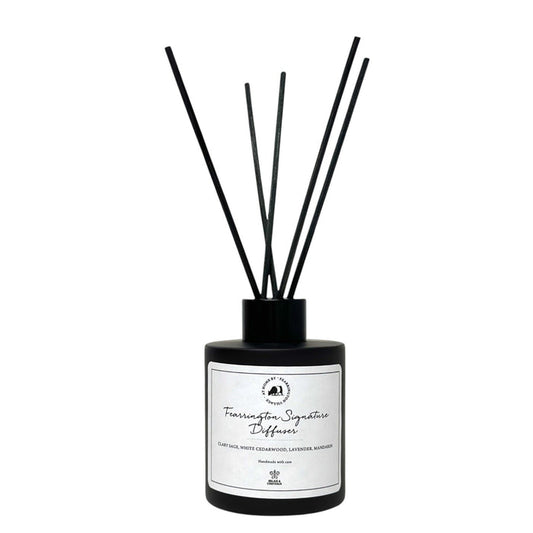 Housed in a sleek black bottle with five slender black reeds, the FEARRINGTON SIGNATURE DIFFUSER from the FEARRINGTON SIGNATURE COLLECTION by FEARRINGTON VILLAGE - HOME DECOR features a white label displaying "Fragrance Signature Diffuser" and scent notes of cedarwood, lavender, and amber. Inspired by the tranquil allure of Fearrington Village, this sophisticated reed diffuser is ideal for anyone who appreciates the calming atmosphere created by a lavender candle.
